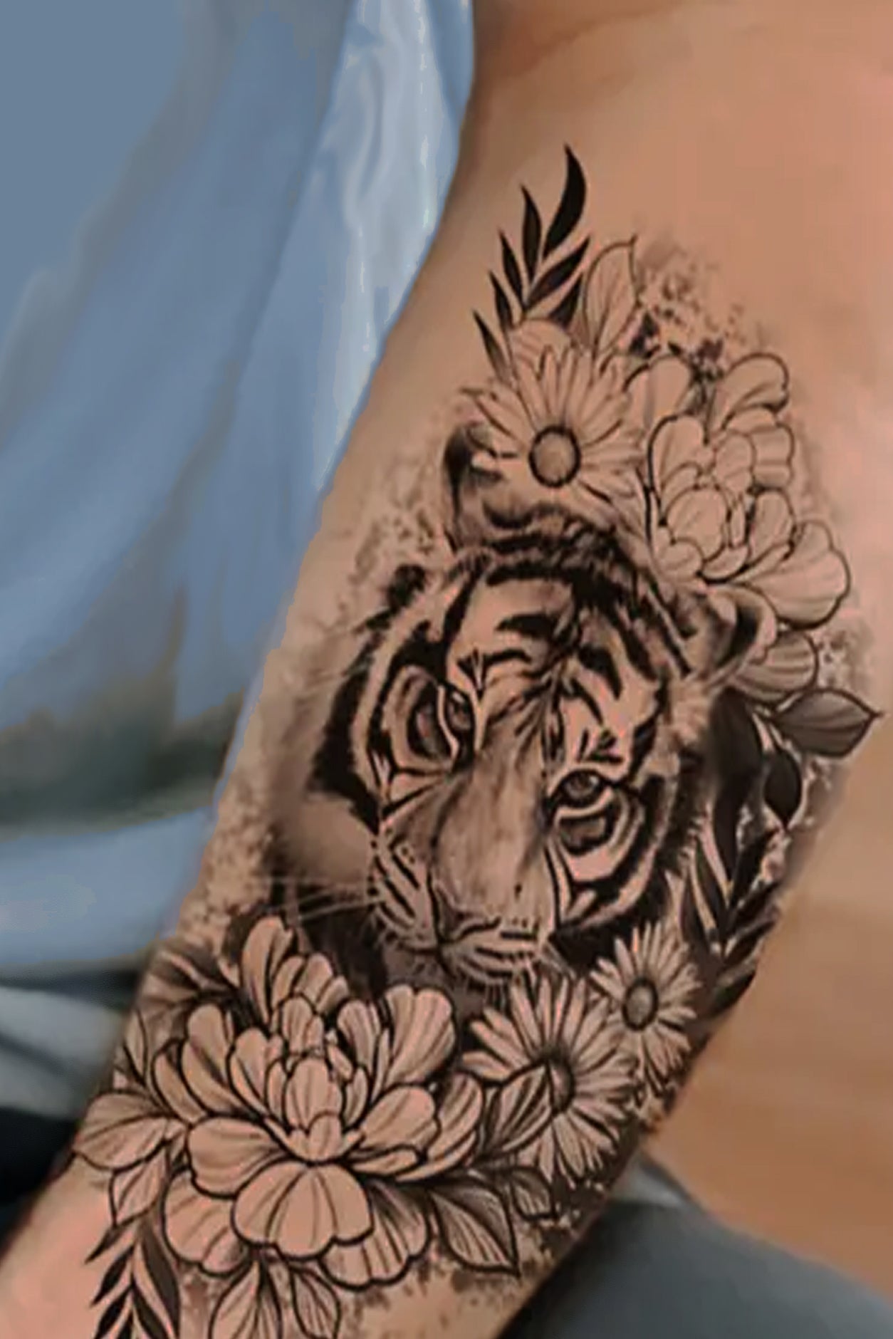 A gender neutral arm displays the tiger surrounded by daisy flowers and peonies, in a black ink realistic tattoo.